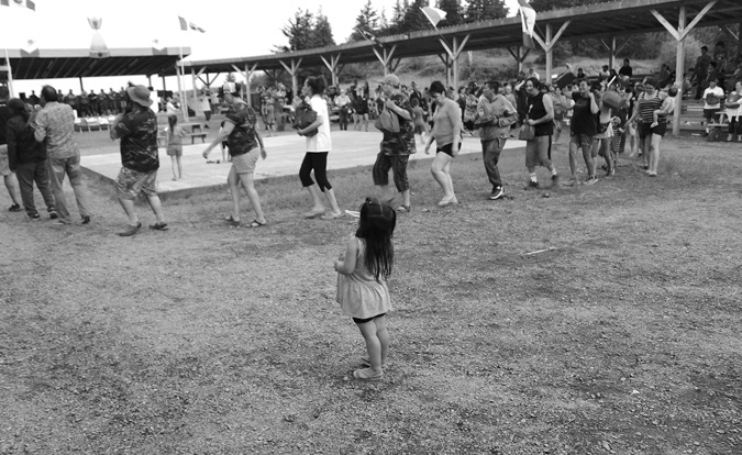 Black and white photograph: A small child is watching people of all ages dance in a circle. The child has long black hair partially held up in pigtails and is wearing a dress, shorts and sandals. In the center of the circle are children on a square stage. On the opposite side of the circle are many people sitting on bleachers under a large wooden canopy. There are two flags on each side of the canopy blowing in the wind.
