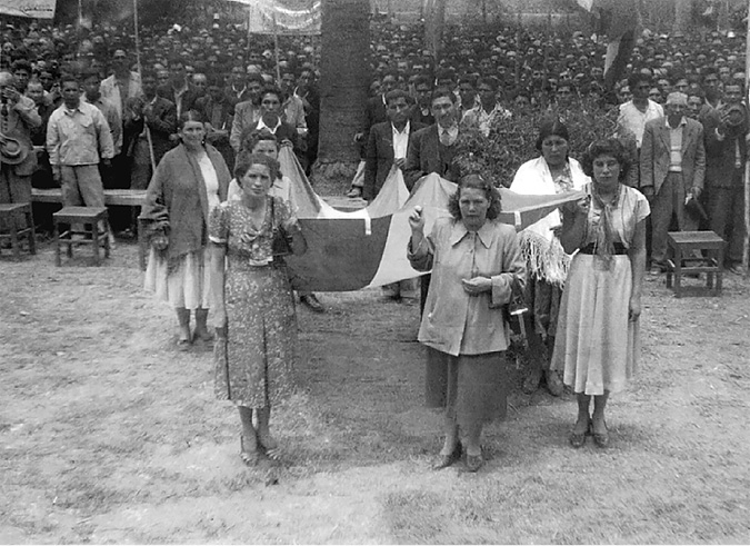 A group of town women hold together a long Bolivian flag and stand in front of a crowd of MNR male militants in an outdoor meeting.