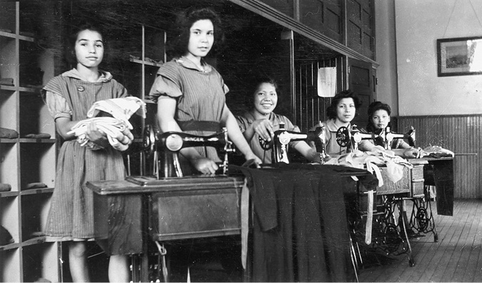 Black and white photograph: Group of five Indigenous female students working in a sewing room using sewing machines.  The two students on the left are standing, and the rest are sitting. Most appear to be teenagers.  They are wearing the same basic work clothing which consists of a basic dress with a short-sleeved shirt underneath.
