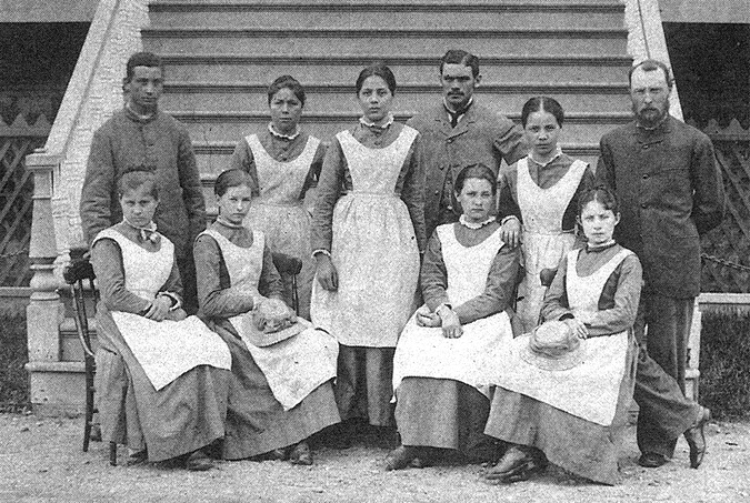 Black and white photograph: Formal photograph of Mohawk Institute staff, consisting of seven women and three men, pictured in front of the steps of the school. The women are wearing the same dress with an apron-like smock over top. Some of the women are seated, while others are standing.  The men are standing behind the women.