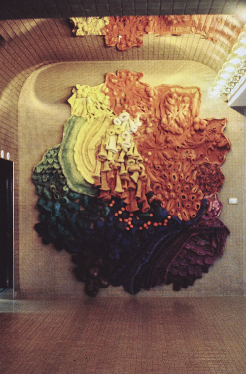 Photo: a textured 3d art mounted on a wall with a row of lights to the right. The art is yellow and orange transitional to green, blue, purple. It has trumpet shapes hanging down, round sections with holes, and is an amorphous shape.