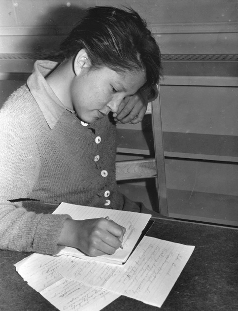 Black and white photograph: Indigenous female student seated at a table writing poetry.  One hand holds a pencil poised to write on the paper below, while the other hand and arm are propped over the back of the chair.