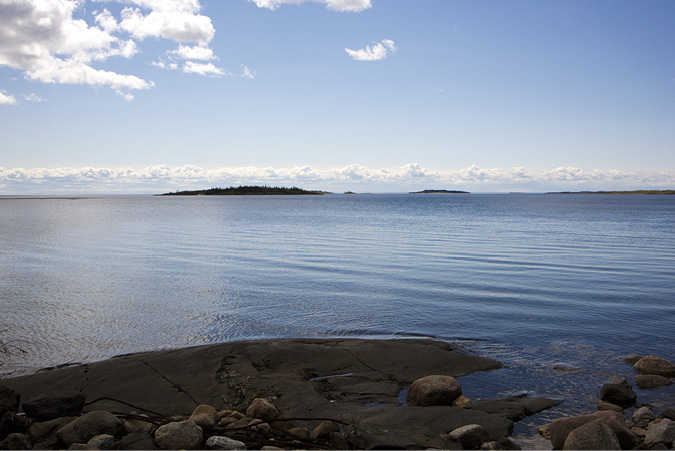 Small rocks sit on the shore of Lake Athabasca that stretches out with small ripples. Far in the distance at the other side of the lake are tree covered hills.