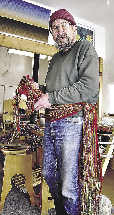 Photo: a man with a scraggly beard and glasses, wearing a red cap, and a red patterned sash on top of jeans and a grey sweatshirt in a workshop