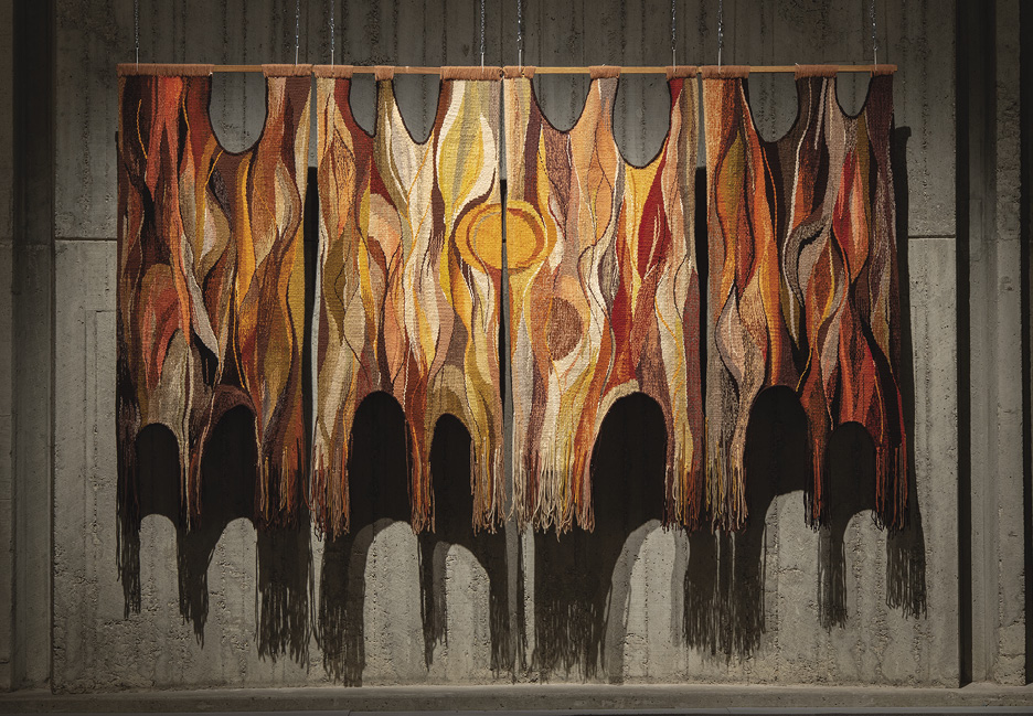 Photo: Four-panelled wall hanging. It is in organize browns, oranges, and yellow. There are “S” shapes throughout, and one notable orange circle in the middle, in addition to semi-circle chunks taken out of the piece along the top and bottom. It hangs on a grey cement wall. 