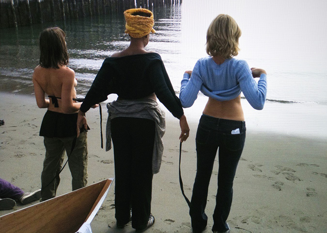 At the same beach, in from of the pointed end of the white boat, 3 people dressed in pants are standing facing the water, only their back bodies are visible as they lift their shirts and bare their front bodies to the water in front of them.