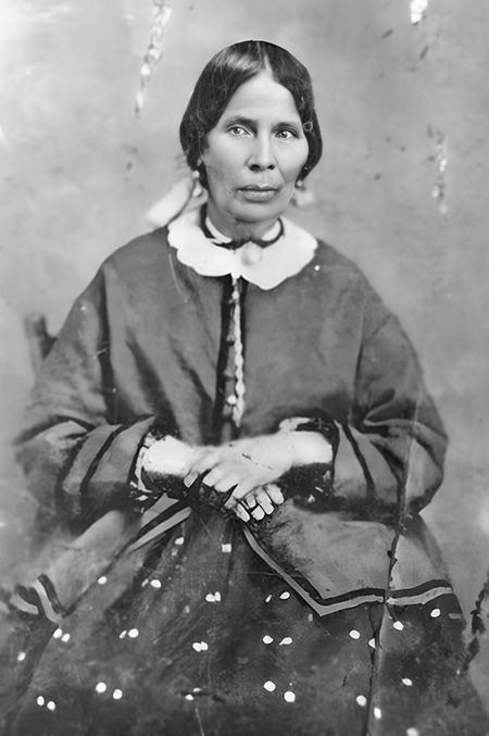 Black and white historical photograph. Portrait of a seated Indigenous woman. She is wearing a dark jacket over a dress with white dots and a white collar. Her black hair is tied neatly back, and she is wearing long white tassel earrings. Her hands are folded in her lap and she has a somewhat startled expression.