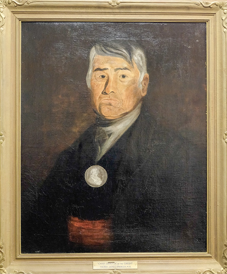Colour painting. Portrait of an Indigenous man in an decorated gold frame. He has short grey hair and a serious expression. He is wearing a black coat, a large silver medal, and a red sash.