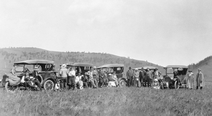 Figure 7-2 “Caravan of automobiles en route to Turner Valley, Alberta.” Black and white photograph. Five early-model automobiles and at least 25 men, women, and children dressed in suits, dresses with some wearing hats pose for a picture as they drive out to see the wells at Turner Valley.