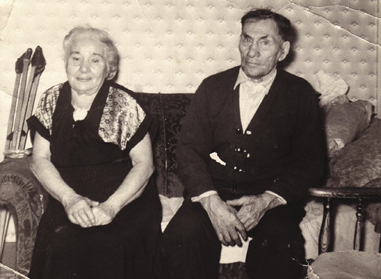 Black and white Photo: an elderly couple sit on a small sofa in front of a wallpapered wall.