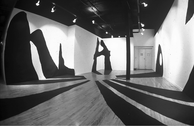 Black and white photograph: The Studio Gallery Nine gallery space, with large, abstract, shadow-like cut outs laid across the floor and adhered the walls. The shadow in the centre of the image, on the far wall, looks like the letter H.
