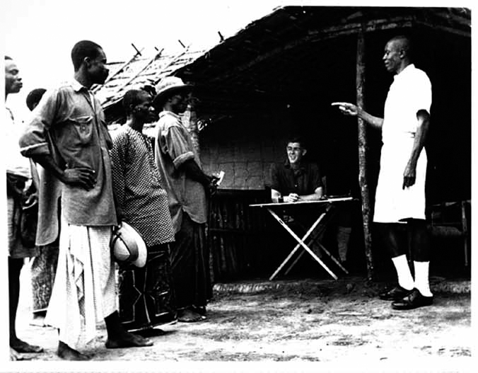A black and white photograph shows a person sitting at a table inside a tent and another person in white attire is talking to the villagers gathered around the tent.