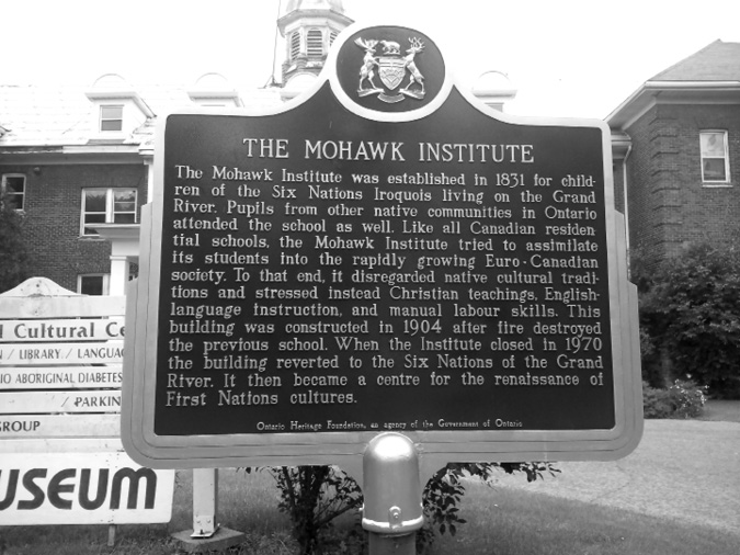 Black and white photograph: Plaque commemorating the Mohawk Institute, located in front of the school. It is metal and the coat of arms for Ontario is at the top of the plaque. The text along the bottom of the plaque reads “Ontario Heritage Foundation, an agency of the Government of Ontario.”