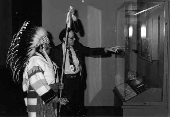 A photograph shows two men, one is wearing a traditional dress with a stick in his hand while the other one is in formal dress. A wooden Almira with a glass covering can be seen.