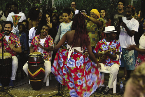 Colour photograph: People dance, clap, and play drums in an open courtyard meeting.