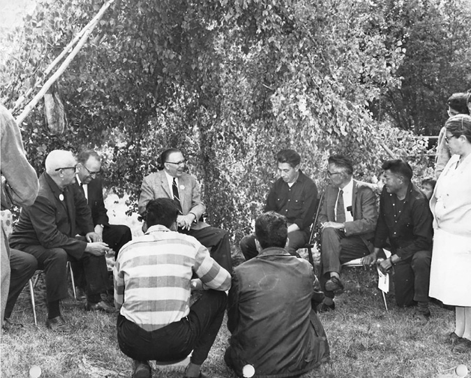 The black and white photograph shows a group of men and one woman gathered in an outdoor setting. The central focus is a group of eight men sitting in a circle, with some casually dressed in suits and others in more informal attire, including shirts and jackets. They appear to be engaged in conversation. In the foreground, two men are crouching, revealing their backs to the camera. A woman, wearing a light-colored dress and glasses, stands to the right, observing the group. Behind them, lush foliage adds a natural backdrop, indicating that they are in a garden or park. The presence of chairs suggests it might be a casual meeting or discussion.