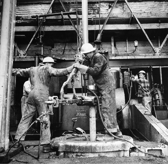 These were workers at a Royalite Company oil site in the Turner Valley. Royalite was affiliated with Imperial Oil from 1921 to 1948 when it became a separate company. The photo shows four rig workers in hard hats and overalls operating a drill while mud seeps around the well head.
