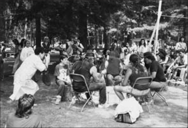 Men, women and children are gathered in the middle of a forest land and talking among themselves. Two men are clicking photographs.