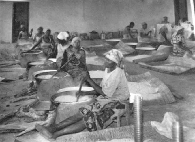 A photograph of women preparing gari in a huge vessel using wood fire inside a building.
