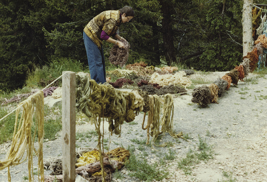Photo: a woman bends down over piles of material on the ground outside behind piles of wool hanging on a strung rope.