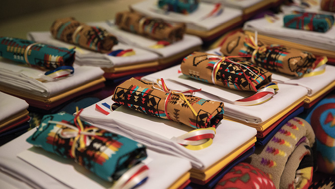 Colour Photograph: Traditional gifts of tobacco wrapped in Indigenous patterned fabric sitting on top of a bundle of coloured broad cloth wrapped in red, yellow, blue and white ribbons.
