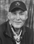 Black and white photograph: Portrait of a smiling man wearing a ball cap, collared shirt, and collared outdoors jacket.