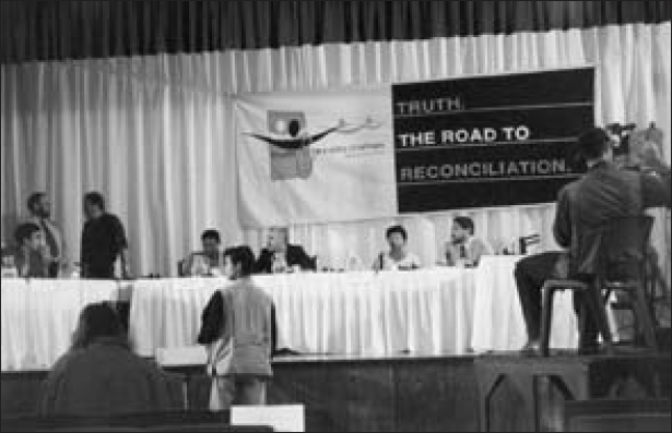A panel of five members is seated in front of a table. Two men are discussing behind them, while another man tapes the meeting on a video camera. The banner above the table contains the logo of the TRC and reads the text, Truth The Road to Reconciliation.