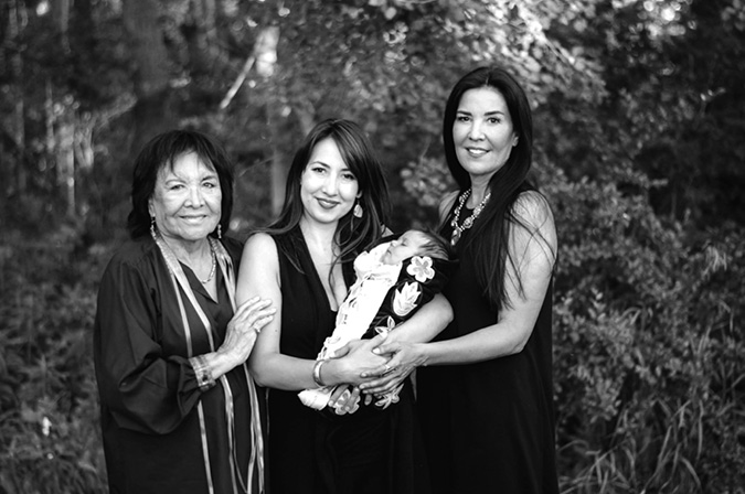 The black and white image depicts three women standing closely together in an outdoor setting with dense foliage in the background. On the left, an elderly woman (Lina Gallup) with short dark hair and earrings wears a dark, long-sleeved top embroidered with ribbons around the collar and sleeve cuff. She stands with her left arm gently resting on the shoulder of the second woman (Billie Fortier), who has long dark hair and earrings, smiles while holding a baby (Soleil Cree Fortier) in her arms. The baby is nestled in a moss bag embroidered with traditional flower beadwork and appears to be asleep. On the right, another woman (Holly  Fortier) with long, straight dark hair also wears a dark dress and a beaded statement necklace, and she smiles at the camera.