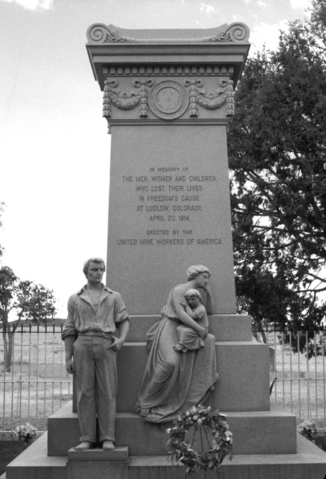 A black and white photo of the granite monument shows a square stone pillar rising from three graduated square stone pedestals. The pillar is topped with stone garlands that frame the seal of the United Mine Workers of America topped by an ornate slightly wider thin top-piece, with a curved scroll-like top. Three carved figures are placed on the pedestals: a miner stands on a thin stone box on the left of the first pedestal, a woman stands on the second pedestal, holding a child, leaning against the monument and facing to the right in the photo, leaning away from the man. He has longish hair, is wearing work pants and a work shirt with the sleeves rolled halfway up his arms. His left hand is crooked, with his thumb in his waistband. The woman wears a long dress. Its skirt falls in folds. Her left elbow rests on the top pedestal, and she leans her head in her left hand. With her right hand, she holds a child propped on her left knee, which rests on a pedestal. The child rests its head and arm against its mother’s arm. The face of the monument reads: In Memory of THE MEN, WOMEN AND CHILDREN, WHO LOST THEIR LIVES IN FREEDOM’S CAUSE AT LUDLOW, COLORADO APRIL 20, 914 ERECTED BY THE UNITED MINE WORKERS OF AMERICA