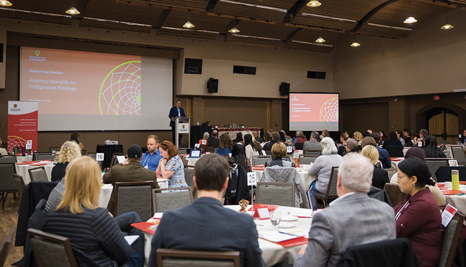 Colour Photograph: Group of people gathered around tables having conversation, with a slide and speaker at the front of the room.