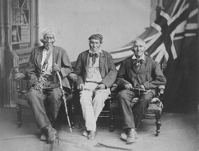 Black and white historical photograph. Three elderly men are sitting on chairs. They are wearing formal dress, and are holding from left to right a cane, a knife, and a tomahawk. There is a bookshelf and a Union Flag in the background.
