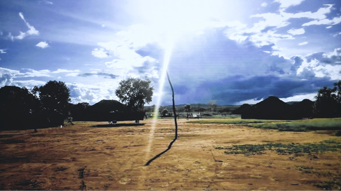 The shadow images of large round hut dwellings in circular village form of the Etenhiritipá Village are located along the basin with sparse trees scattered at the edges. The sun is beaming down on the village with only a few low clouds braking the heat.