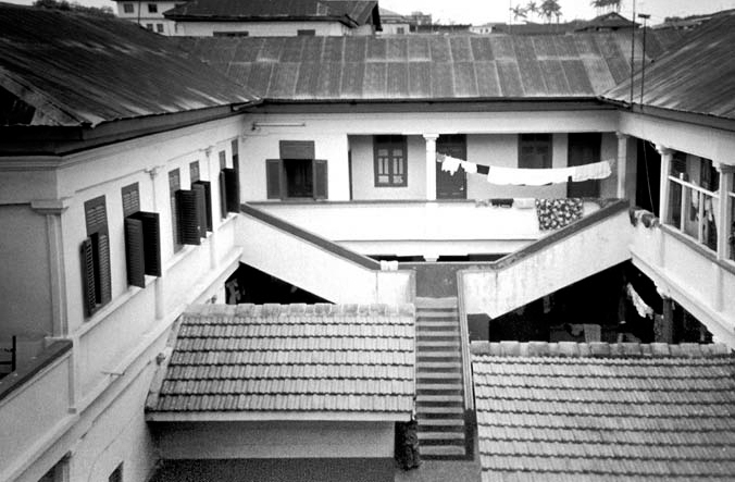 A black and white photograph of the family house of President Kufuor in Kumasi, Ghana. A white building with black slanted roofs, surrounding a courtyard.