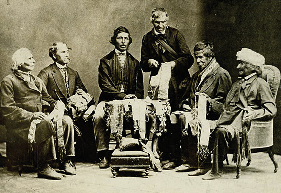 Black and white historical photograph. Six men, five of them Indigenous, are in a semi-circle surrounding a small round table covered in wampum belts. Five of the men are seated, and one is standing in the middle looking over them. The men are all formally dressed, mature or elderly, and holding an assortment of wampum belts. The standing man is pointing at his and explaining.