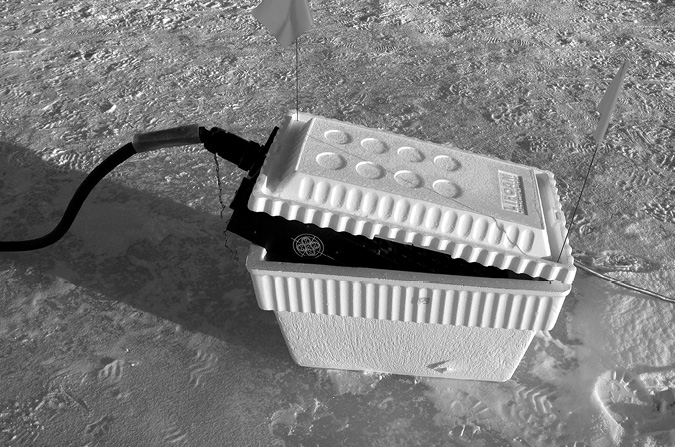 Black and white photograph, a white polystyrene ice cooler sits on a frozen lake; a generator sits inside with cords running off to both sides of the image; there are two small white flags stuck into the cooler.