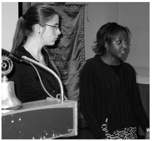 A black and white photograph of Cayla Gilbert watching as Eunice Mahomah speaks on the stage of Calgary Olympic Park Rotary Club, May 2005.