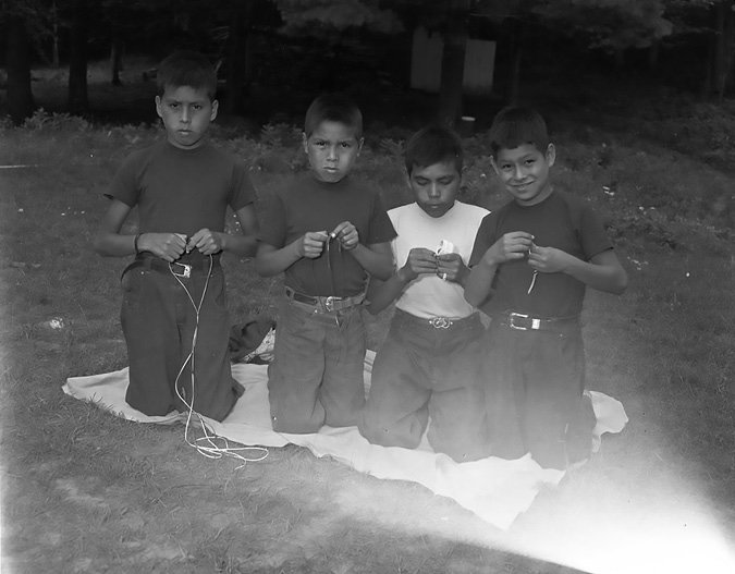 Black and white photograph: Four Indigenous boys outdoors on their knees who are dressed casually in pants and t-shirts.  All four are kneeling on a blanket and working on some sort of craft.