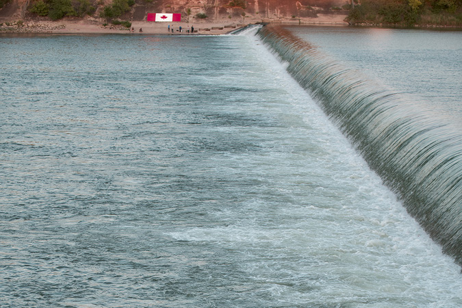 This image was created on the shore of the South Saskatchewan River in Saskatoon on Canada Day. The photo affords a view across the wide expanse of the river where the city weir is located; on the far bank are a half-dozen people lounging on a narrow strand and fishing in the churning water; behind the human figures a large Canadian flag is emblazoned across the rising riverbank.