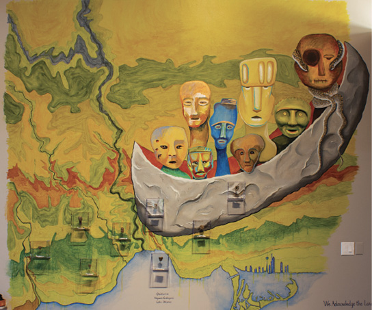Colour Photograph. Colourful mural, showing a green and yellow landscape with rivers running through it and a lake at the bottom. A large grey canoe is filled with spirit figures, with stylized faces of varying colours.