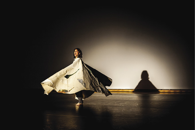 Photo: a person in a dark room under spotlight whirling with their mouth open while twirling with a large white and blue cloak on. 