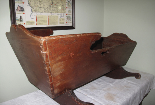 Colour photograph. A worn-looking wooden cradle sitting on a table, with a map behind it. The cradle has a high back, handles on the side, and carved feet.