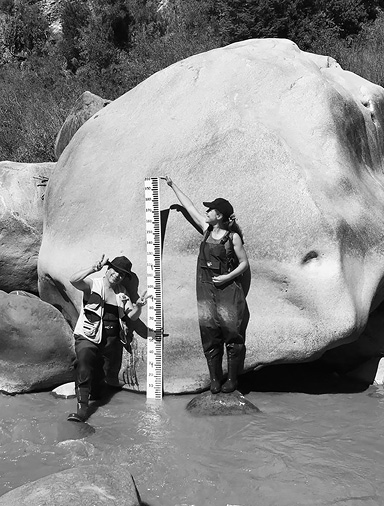Black and white photograph: Photo of two community researchers in hip waders standing in the river and on a rock, with a large measuring board taller than they are, marked off with height measurements.