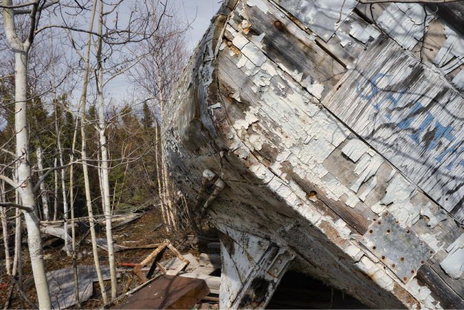 A long abandoned wooden boat sits on a shoreline, Paint is chipping off the structure. Bare trees surround the structure.