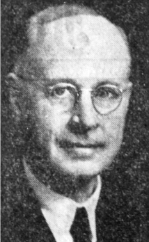 Black and white photograph: A portrait of an older man wearing round glasses and a suit with a tie. He has short hair and a serious expression on his face.