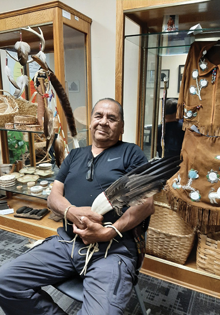 Colour photograph. Photograph of an Indigenous man seated at a gallery display. He is wearing a navy shirt and pants. He is smiling, and holding a fan of eagle feathers. A variety of First Nations objects, including a buckskin dress and woven baskets, are displayed on tables and in cabinets behind him.