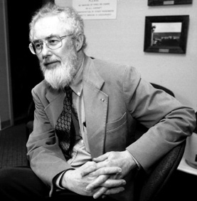 Black and white photograph: A bearded elderly man wearing glasses and a suit jacket and tie sits in a chair with hands clasped. Behind him are a few framed pictures on a wall.