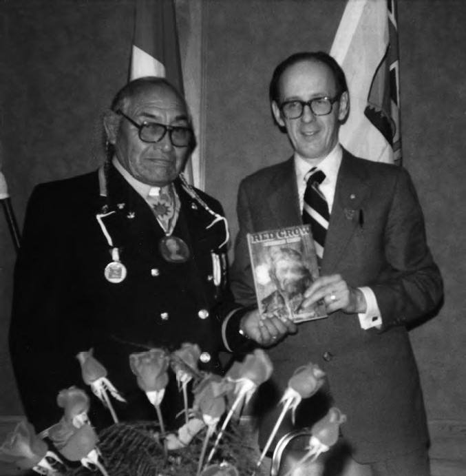 A photograph shows two men in formal dress, holding a book in their hands. Two flags can be seen in the background.