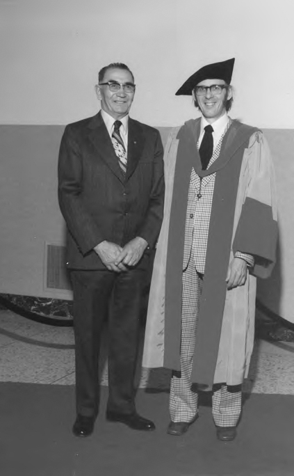 A photograph of two people wearing formal dress with spectacles. The man on the left is wearing a plain dark colour coat while the other is wearing a long overcoat with a light checked coat. The background has a wall with different shades on it.