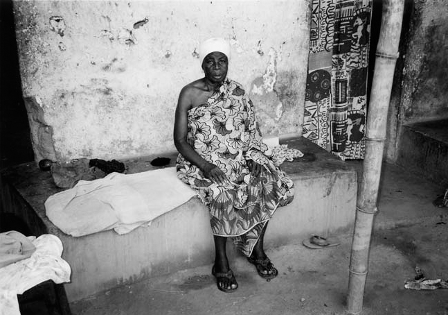A black and white photograph of Obaapanyin Yaa Kronama, Queenmother of Anyinam sitting on a slab outside the house wearing an African dress.