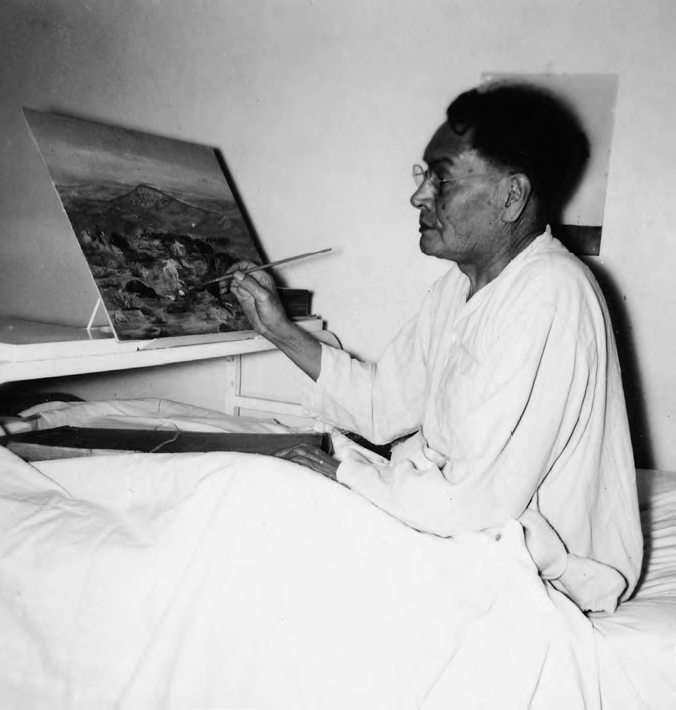 A photograph shows a man wearing spectacles and sitting on a bed with one painting. He has a paintbrush in his hand.